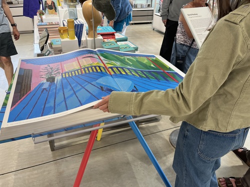 Millie turning the pages of the giant Hockney SUMO book, revealing a vivid blue and pink poolside painting across the double-page spread