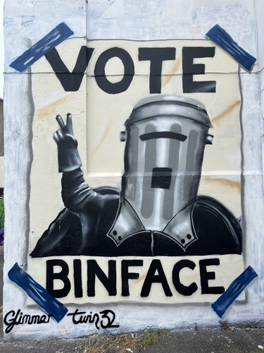 A large street art mural on the side of a building in Brighton reading Vote Binface, showing the comedy political candidate with a bin for a head giving a peace sign, by Glimmer Twin 32