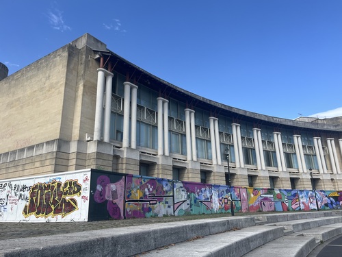 The curved concrete facade of a former Lloyd's Bank building, with a colourful mural along its graffitied hoarding