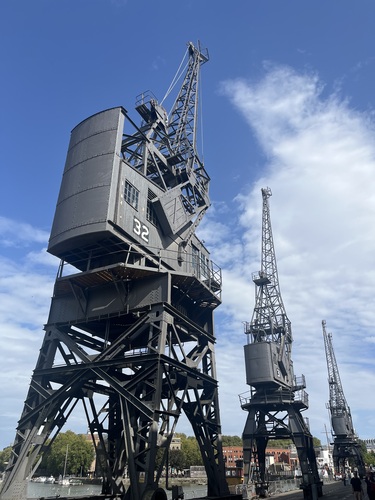 Tall black dockside cranes with angular, futuristic silhouettes against a blue sky with light cloud