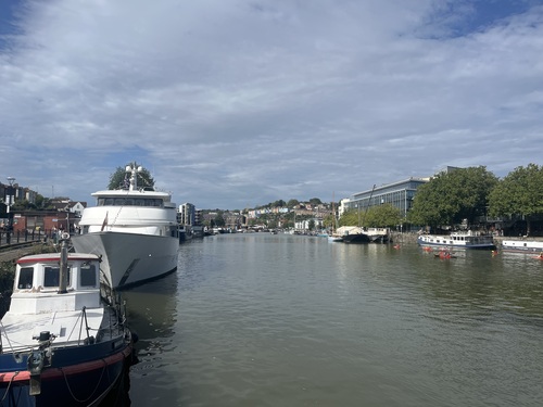 Boats moored along Bristol's floating harbour, with kayakers paddling past and colourful houses on the hillside beyond