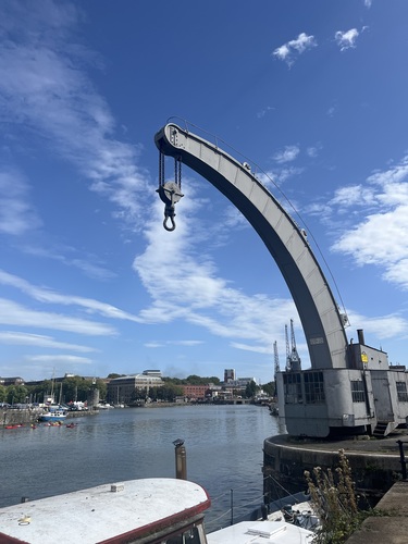 A curved grey dockside crane arm with hook and chains hanging over the harbour