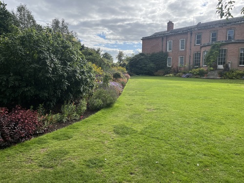 A wide lawn bordered by a flowering shrub border, with Dunham Massey's red brick house visible in the background
