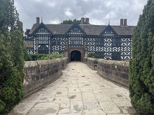 Black and white timber-framed Speke Hall seen from the approach path over the old moat, with a tree either side