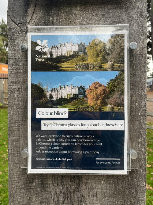 Poster at Sheffield Park headed 'Colour blind?', advising that EnChroma glasses for colour blindness can be borrowed free from reception. Two photos of the house and gardens show the scene before and after colour correction.